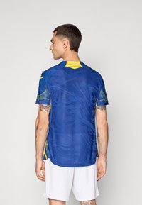 Maglia sportiva blu con un motivo testurizzato, caratterizzata da accenti gialli e un logo ricamato sulla schiena. Maniche corte, tessuto leggero.