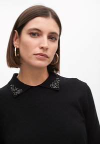 Černý pletený svetr s kulatým límcem, zdobeným jemnou černou výšivkou. Modelka má krátké vlasy a zlaté kruhové náušnice.