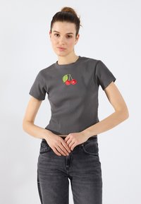 Grå stripete t-skjorte med kort erme som har et brodert kirsebærdesign i rødt og grønt i midten. Brukt med mørk grå jeans.
