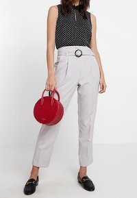 Černý top bez rukávů s puntíky, šedé kalhoty s vysokým pasem a kulatou sponou, drží červenou kulatou kabelku, černé loafers.