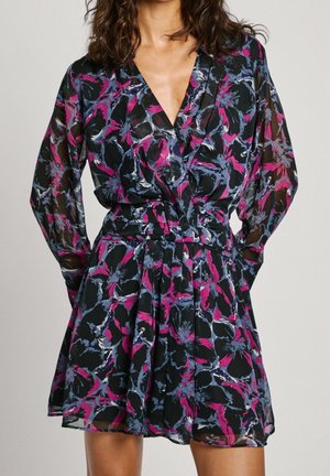 Vrouw draagt een doorschijnende, korte jurk met lange mouwen, een roze, blauwe en zwarte abstracte bloempatroon en een V-hals.