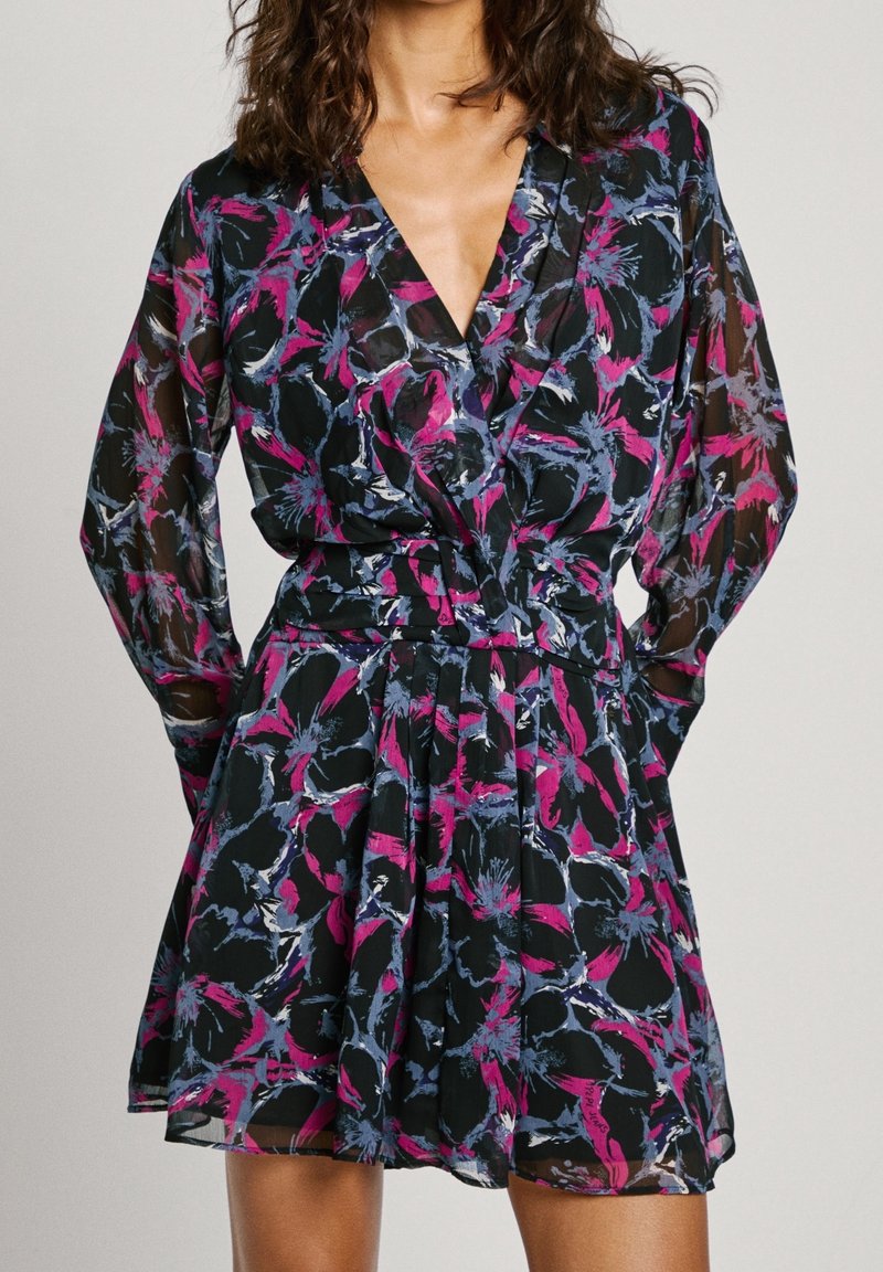 Femme portant une robe courte transparente à manches longues avec un motif floral abstrait rose, bleu et noir et un col en V.