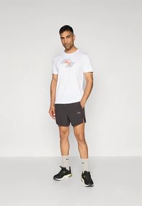 Mees seisab ühe käe taskus, seljas valge Puma T-särk, mustad lühikesed püksid, beeži värvi Puma sokid ja mustad spordijalatsid, neutraalse heledal taustal.