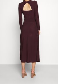 Robe longue bordeaux à manches longues avec texture côtelée, corsage ajusté, jupe évasée en A et ouverture en trou de serrure au dos. Portée avec des sandales à talons carrés couleur beige.