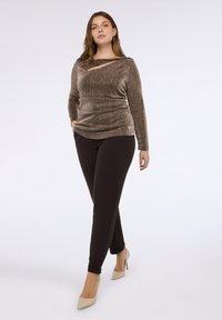 Błyszcząca brązowa bluzka z długim rękawem z ukośnym wycięciem; w połączeniu z czarnymi dopasowanymi spodniami i beżowymi szpilkami, stoi na białym tle.