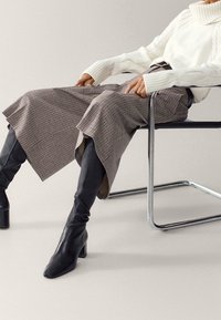 Personne assise, portant un pull à col roulé blanc, un pantalon large à carreaux et des bottes noires montantes à talons, sur une chaise à armature métallique.