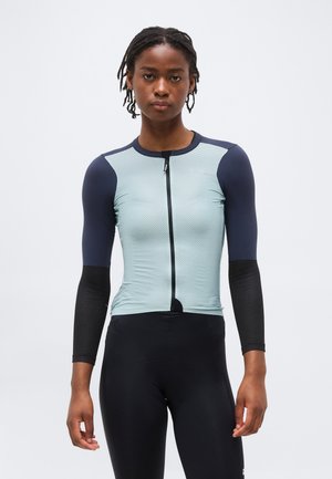 Γυναίκα με κοντά πλεγμένα μαλλιά, φοράει μακρυμάνικη μπλούζα ποδηλασίας σε ανοιχτό μπλε και μαύρο χρώμα και μαύρο παντελόνι ποδηλασίας, στέκεται όρθια.