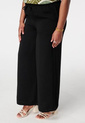 Femme portant un pantalon large noir avec une bordure en dentelle, des sandales blanches à talons blocs à brides, et des bracelets dorés à un poignet.