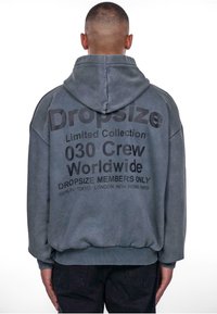 Kapuzensweatshirt in gedecktem Grau mit auffälligem schwarzen Text auf der Rückseite. Design mit fallenden Schultern und gerippten Bündchen und Saum.