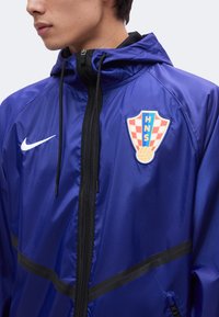 Άτομο που φοράει μπλε μπουφάν Nike με κουκούλα, μπροστινό φερμουάρ και κόκκινο-άσπρο καρό λογότυπο αθλητικού συλλόγου HNS στο στήθος.