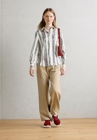 Chemise à rayures, à manches longues, en blanc et bleu marine ; pantalon large en kaki ; baskets rouges. Le modèle porte un petit sac à main rouge. Fond en bois.