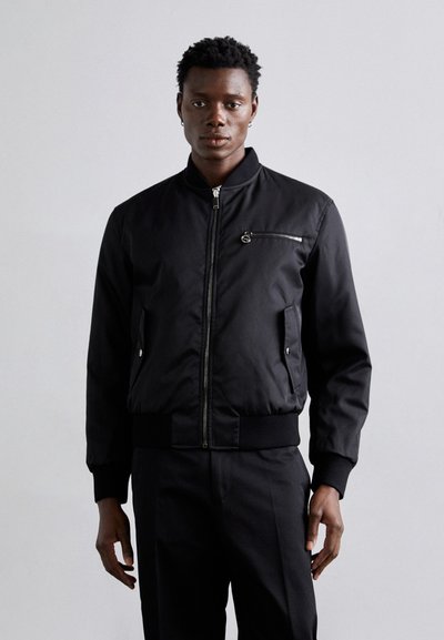 Veste bomber noire en tissu lisse avec une fermeture éclair à l'avant, des poignets côtelés et une poche zippée sur la poitrine. Design ajusté.