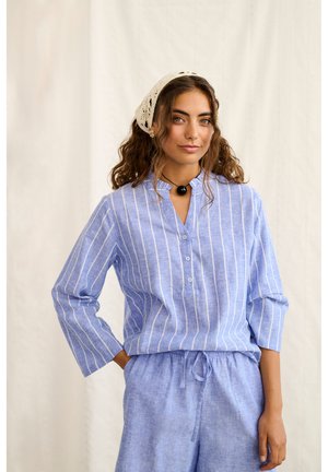 Lokkis juustega naine kannab sinist triibulist pika varrukaga särki ning sobiva nööriga pükse, beeži peapaela ja musta ripatsiga kaelakeed.