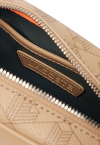 Καφέ δερμάτινη θήκη με υφή και μεταλλικό φερμουάρ. Το εσωτερικό διαθέτει σκουρόχρωμη υφασμάτινη επένδυση και πορτοκαλί διακοσμητική λεπτομέρεια. Ετικέτα Lacoste στο εσωτερικό.