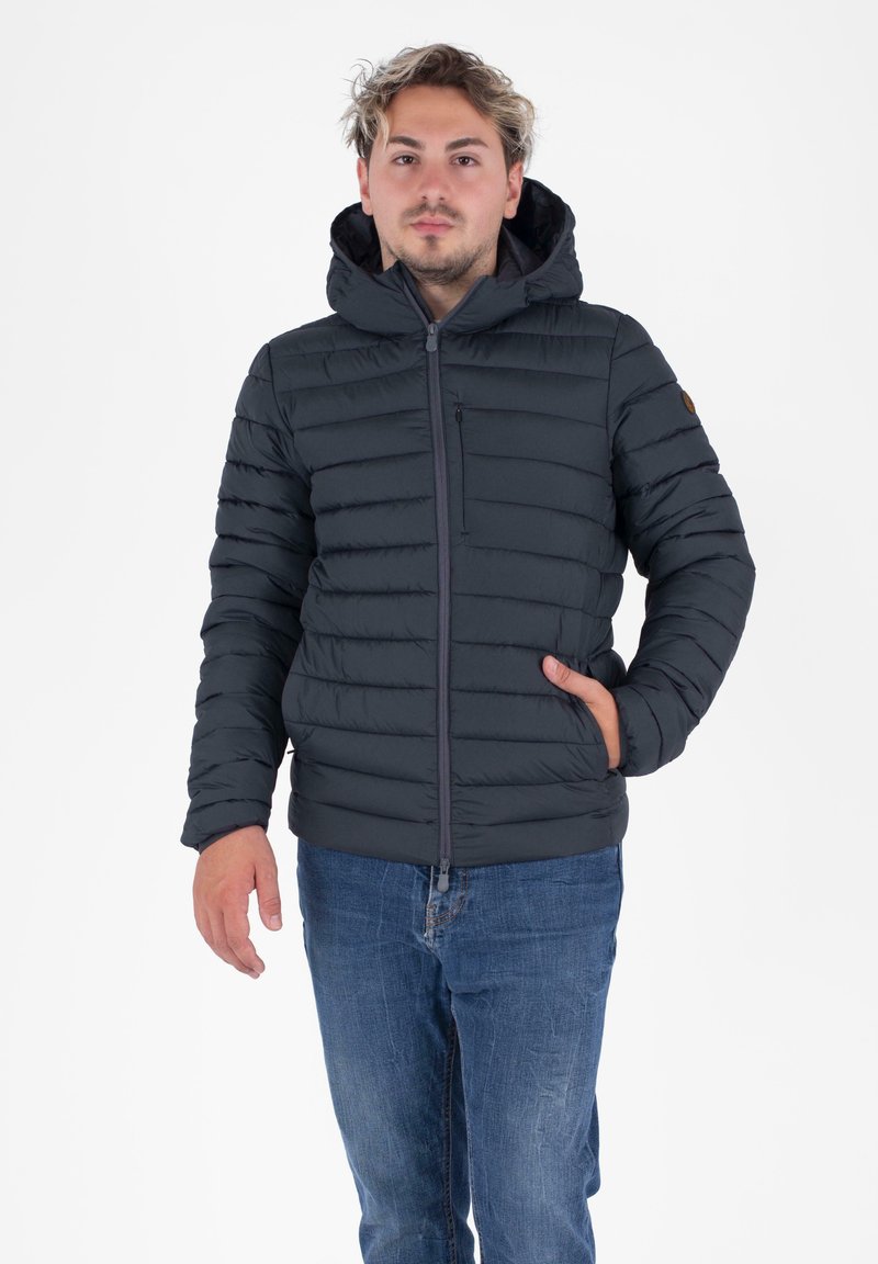 Giovane uomo che indossa un giubbotto con cappuccio imbottito nero e cerniera, jeans blu, con una mano nella tasca del giubbotto, in piedi su uno sfondo bianco.