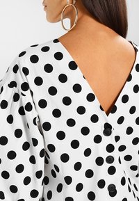Weiße Bluse mit schwarzen Punkten, V-förmiger Rücken, geraffte Ärmel und schwarze Knöpfe entlang des Rückens. Glatte Textur.