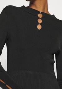 Haut noir côtelé à manches longues avec un col montant et trois découpes ovales ornées d'accents métalliques à l'avant. Tissu texturé avec une coupe ajustée.