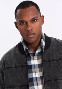 Mustat pommikavereet, joissa on teksturoitu pinta, sinisen ja ruskean ruudullisen paidan päällä. Kaulus on ribattu, ja siinä on napit ja vetoketjukiinnitys.