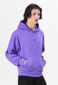Lila Kapuzenpullover mit einer frontalen Tasche, Kordelzug-Kapuze und Logodetail. Hergestellt aus weichem Stoff mit gerippten Säumen und einem lockeren Schnitt.