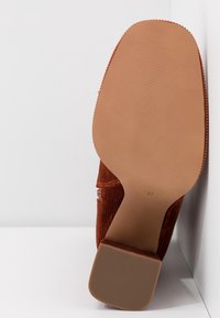 Semelle d'une bottine en daim couleur rouille avec un talon large, taille 37, placée debout contre un mur d'angle blanc.