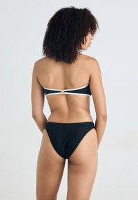 Black bikini set featuring a bandeau top with a gold clasp and white accents, paired with a textured high-waisted bottom.
