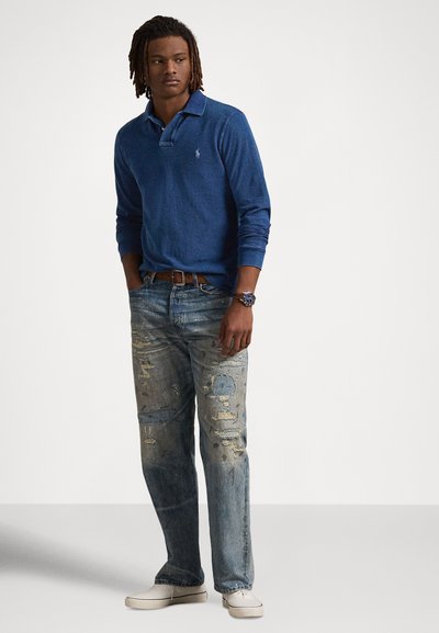 Μπλε πόλο με μακριά μανίκια, υφασμάτινο με υφή, με ανοιχτόχρωμα τζιν με φθορές και μπεζ παπούτσια slip-on, φορώντας ρολόι και καφέ ζώνη.