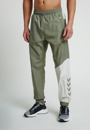 Mężczyzna ubrany w sportowe spodnie w blokach kolorystycznych oliwkowej zieleni i bieli oraz czarno-szare sneakersy, stoi z rękami swobodnie opuszczonymi wzdłuż ciała.