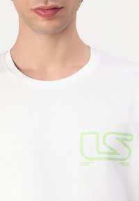 Hvit bomulls T-skjorte med rund hals og en neongrønn "LS" logo på venstre bryst. Glatt tekstur med minimale designdetaljer.