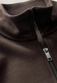 Nærbillede af brun stof med en delvist lynet sort metallynlås og en opretstående krave.