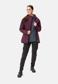 Giacca isolante bordeaux con zip, indossata sopra un top grigio, abbinata a pantaloni neri e calzature robuste nere.