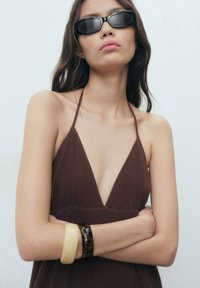 Robe halter marron avec un décolleté en V profond. Le modèle porte deux bracelets : un crème et un en écaille sombre. Des lunettes de soleil noires complètent le look.