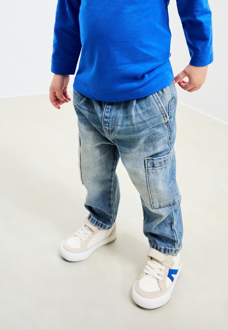Chemise bleue à manches longues, pantalon cargo en denim bleu clair avec taille élastique, et baskets blanches avec des accents beige et bleu.