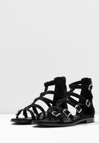 Mustat nupukkinahasta valmistetut sandaalit, joissa on useita ohuita hihnoja, hopeiset solkiyksityiskohdat, avonaiset varpaat ja litteät pohjat. Sopivat lämpimään säähän.