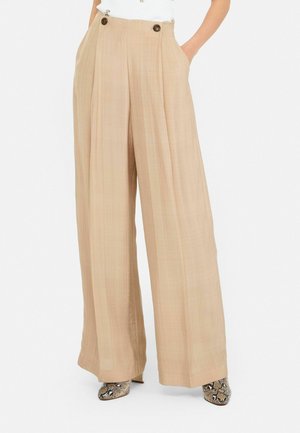 Pantalons larges beige clair avec double bouton à la taille, en tissu texturé et avec poches. Associés à des chaussures à talons hauts à motifs.
