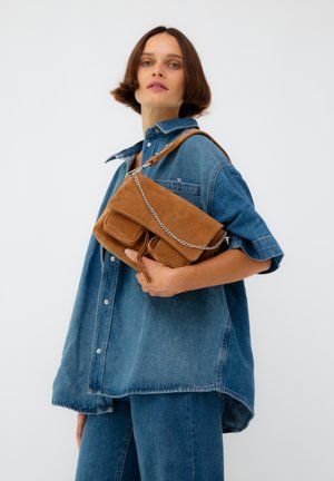Femme portant une chemise en denim oversize et un jean, tenant un sac à main en daim marron avec des détails en chaîne, sur un fond blanc uni.