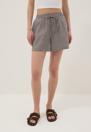 Mujer con pantalones cortos de cuadros en blanco y negro con cordón, combinados con sandalias marrones de doble hebilla y una camiseta corta blanca.
