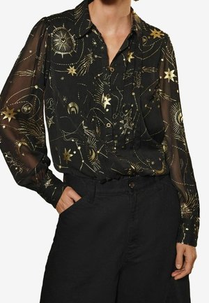 Blouse noire transparente à manches longues avec des imprimés célestes dorés brillants, comprenant des étoiles et des lunes, portée rentrée dans un pantalon noir.