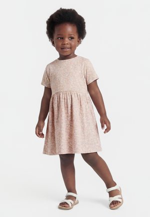 Toddler girl with curly hair wearing a short-sleeve, light pink floral dress and white sandals, standing with one leg forward.