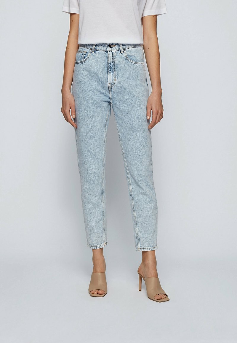 Donna che indossa jeans a vita alta blu chiaro, t-shirt bianca e scarpe con tacco aperte beige, in piedi davanti a uno sfondo semplice.