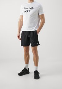 Bílé přiléhavé tričko s černým logem "Reebok", doplněné o černé sportovní šortky. Kompletní outfit tvoří černé tenisky a šedé ponožky.