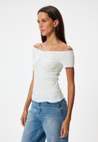 Valkoinen olkapäiltä avonainen paita ribatusta kankaasta ja pitsireunuksella, vartalonmyötäinen malli, yhdistettynä sinisiin farkkuihin.