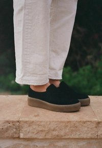 Juodos suėtos slip-on tipo bateliai su apvaliu batu, su šviesiai rudu guminiu padu. Sukomplektuoti su baltais trumpais džinsais.