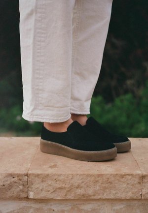 Scarpe slip-on in suede nero con punta rotonda, dotate di una suola in gomma marrone chiaro. Indossate con pantaloni in denim bianchi a vita alta e cropped.