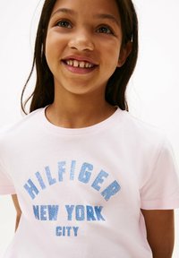 Camiseta de algodón rosa con texto azul brillante que dice "HILFIGER NEW YORK CITY". Diseño de cuello redondo y mangas cortas. Textura suave.