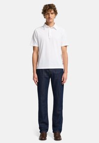 Polo bianco con colletto, abbinato a jeans dritti blu scuri. Il modello indossa scarpe marroni senza lacci, mostrando un outfit casual.