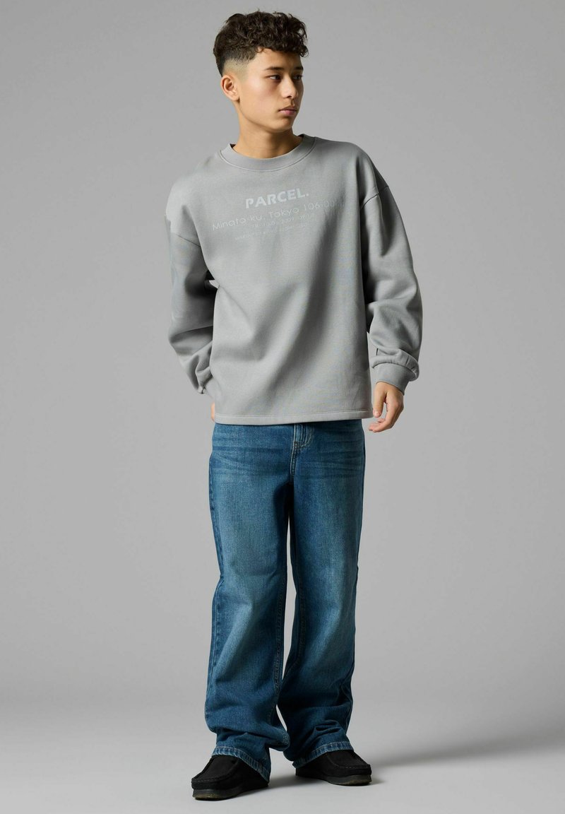 Jeune personne aux cheveux courts et bouclés portant un sweat-shirt gris oversize et un jean bleu ample, debout devant un fond gris uni.