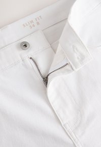 Pantalon blanc slim fit avec fermeture éclair partiellement ouverte et bouton métallique, taille indiquée 34 R à la ceinture.