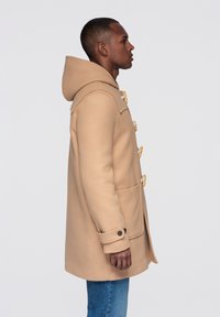 Beige duffelcoat med huva, träknappar och två framfickor. Tillverkad av ett texturerat tyg; har en rak silhuett.