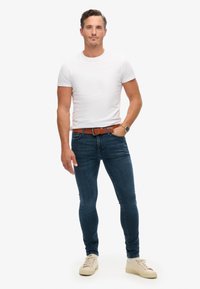 Camiseta de algodón blanca combinada con jeans ajustados de color azul oscuro. Cinturón de cuero marrón con herrajes metálicos. Zapatillas de lona color beige claro con cordones.