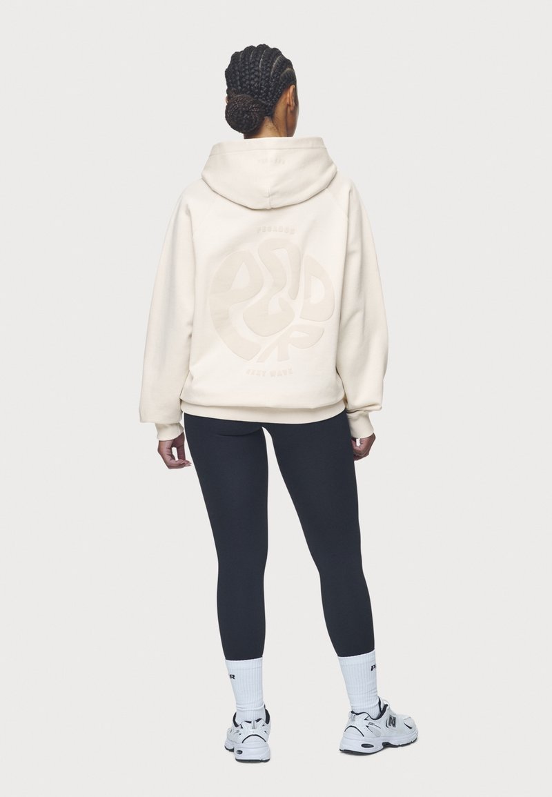 Sudadera con capucha color crema con un gran gráfico tonal en la espalda, combinada con leggings negros y zapatillas deportivas blancas. Pelo trenzado recogido hacia arriba.